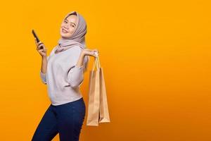 Portrait of cheerful asian woman holding phone and shopping bag over yellow background photo