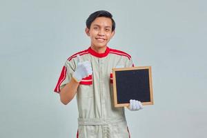 Portrait of handsome young mechanic holding blank board and thumbs up gesture with smiling expression on gray background photo
