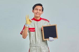 Cheerful young mechanic wearing uniform doing ok gesture and showing blank board on gray background photo