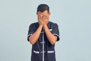 Portrait of frustrated Asian young man wearing taekwondo kimono covering face with palms isolated on gray background photo