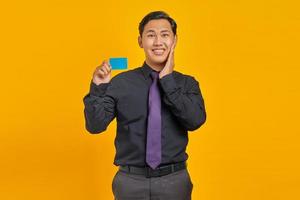 Feliz joven empresario asiático sosteniendo una tarjeta de crédito y tocando la mejilla sobre fondo amarillo foto