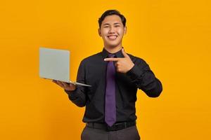 Portrait of smiling young Asian businessman pointing finger at laptop on yellow background photo