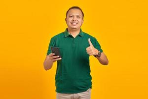 Portrait of cheerful Asian young man holding mobile phone and showing thumbs up sign over yellow background photo