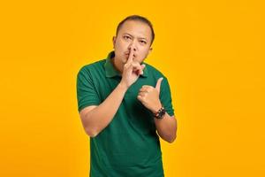 Attractive asian man gesturing to silence and pointing aside over yellow background photo