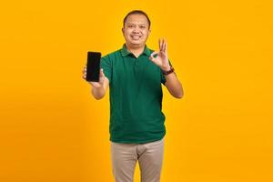 Happy Asian man showing mobile phone blank screen and hand gesture okay on yellow background