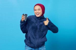 Cheerful beautiful young woman holding coffee cup and showing thumbs up gesture over blue background photo