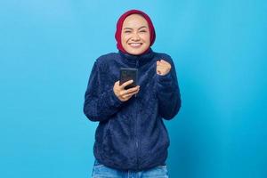 Feliz joven mujer asiática mediante teléfono móvil con éxito gesto de la mano sobre fondo azul.