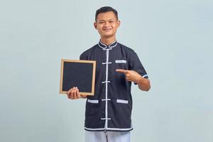 Retrato de alegre joven asiático vistiendo uniforme de karate apuntando a la placa en blanco con el dedo sobre fondo gris foto