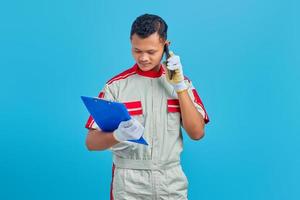 Serious looking young Asian mechanic talking on mobile and checking notes on clipboard isolated on blue background