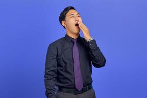 Handsome young businessman feeling sleepy and covering mouth with hands on purple background