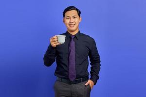 Handsome young businessman smiling at camera with hands in pocket and holding mug on purple background