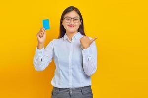 Sonriente joven mujer asiática señalando con el dedo en el espacio de la copia y sosteniendo la tarjeta de crédito sobre fondo amarillo foto