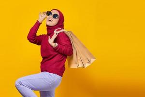 Cheerful young Asian woman wearing sunglasses and holding shopping bags on yellow background photo