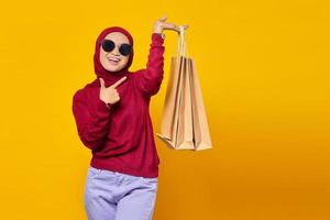 Smiling young Asian woman pointing fingers at shopping bags on yellow background photo