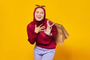 Happy young Asian woman with hand on a chest and holding shopping bags on yellow background