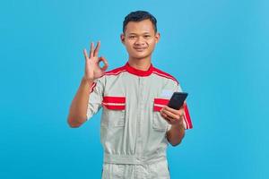 Portrait of smiling young mechanic making okay gesture and showing approval symbol isolated on blue background photo