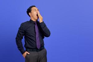 Portrait of handsome young Asian man feeling sleepy and covering mouth with hand on purple background
