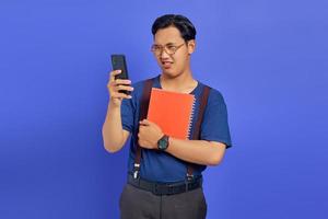 Excited young handsome student looking at cell phone and holding notebook on purple background photo