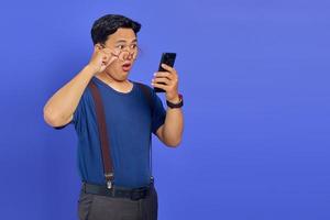 Shocked Asian young man looking at smartphone screen and taking off glasses on purple background photo