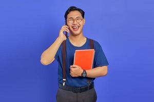 Alegre joven estudiante hablando por teléfono inteligente mientras sostiene el portátil sobre fondo púrpura foto