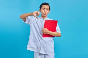 Enfermero joven enojado sosteniendo la carpeta roja y el pulgar hacia abajo gesto sobre fondo azul. foto