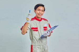 Alegre joven mecánico sosteniendo el portapapeles y apuntando hacia arriba con lápiz sobre fondo gris foto
