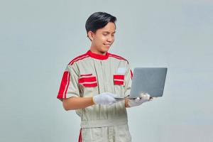 Portrait of smiling handsome mechanic man wearing uniform holding and using laptop on gray background photo