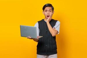 Portrait of surprised young Asian man holding laptop with hand on cheek on yellow background