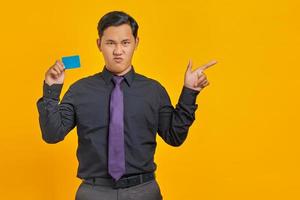 Enojado empresario asiático sosteniendo la tarjeta de crédito con el dedo acusador en el espacio de la copia sobre fondo amarillo foto