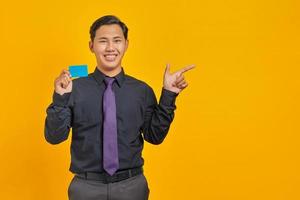 Smiling asian businessman holding a credit card while pointing finger at copy space on yellow photo