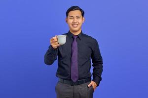 Handsome young businessman smiling at camera with hands in pocket and holding mug on purple background photo