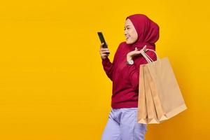 Smiling young Asian woman using a mobile phone and holding shopping bags on yellow background photo