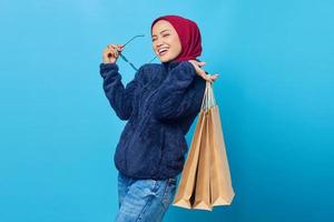 Cheerful young Asian woman biting glasses and holding shopping bag on blue background photo