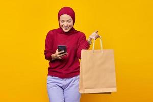 Smiling young Asian woman reading message on smartphone and showing shopping bags on yellow background photo