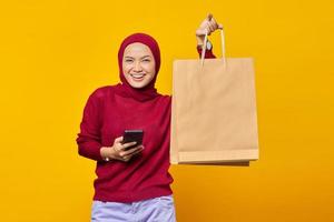 Cheerful young Asian woman holding smartphone and showing shopping bags on yellow background photo