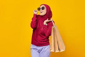 Cheerful young Asian woman wearing sunglasses and holding shopping bags on yellow background photo