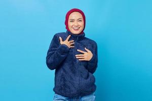 Cheerful young Asian woman put a hand on a chest and looking at camera on blue background photo