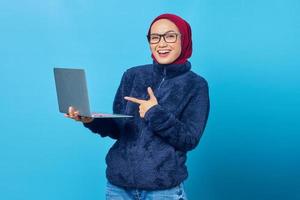 Beautiful asian student wearing blue jacket holding laptop with smiling expression on blue background photo