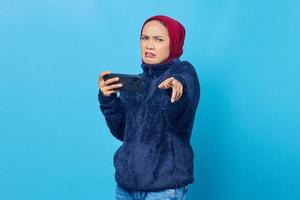 Sad young asian woman playing video game on mobile phone and pointing finger at camera on blue background