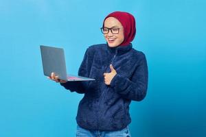 Hermosa estudiante asiática vistiendo chaqueta azul usando laptop y dando pulgar hacia arriba con expresión sonriente