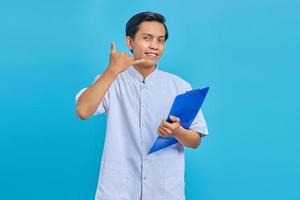 Portrait of smiling male nurse holding clipboard standing and making call gesture on blue background photo