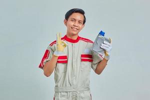 Retrato de hombre guapo alegre vestido con uniforme de mecánico sosteniendo una botella de plástico de aceite de motor y mostrando aprobación con el pulgar hacia arriba sobre fondo gris foto