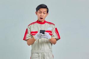 sorprendido, guapo, hombre, llevando, mecánico, uniforme, utilizar, teléfono celular, aislado, en, fondo gris foto