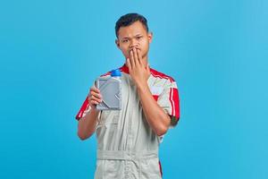 Shocked young Asian mechanic portrait holding engine oil plastic bottle and covering mouth on blue background hands