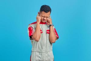 Retrato de frustrado joven mecánico asiático cubriendo las orejas con el dedo sobre fondo azul.