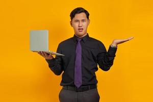 Surprised handsome young businessman showing copy space in palm and holding laptop on yellow background photo