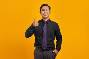Alegre joven empresario asiático mostrando Thumbs up gesto aislado sobre fondo amarillo