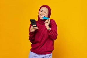 Smiling young Asian woman holding mobile phone and biting credit card on yellow background