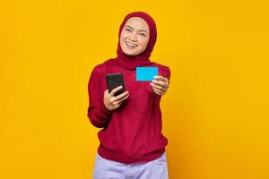 Sonriente joven mujer asiática mostrando tarjeta de crédito y sosteniendo un teléfono móvil sobre fondo amarillo foto