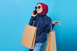 Portrait of smiling young Asian woman talking on mobile and holding shopping bags on blue background photo